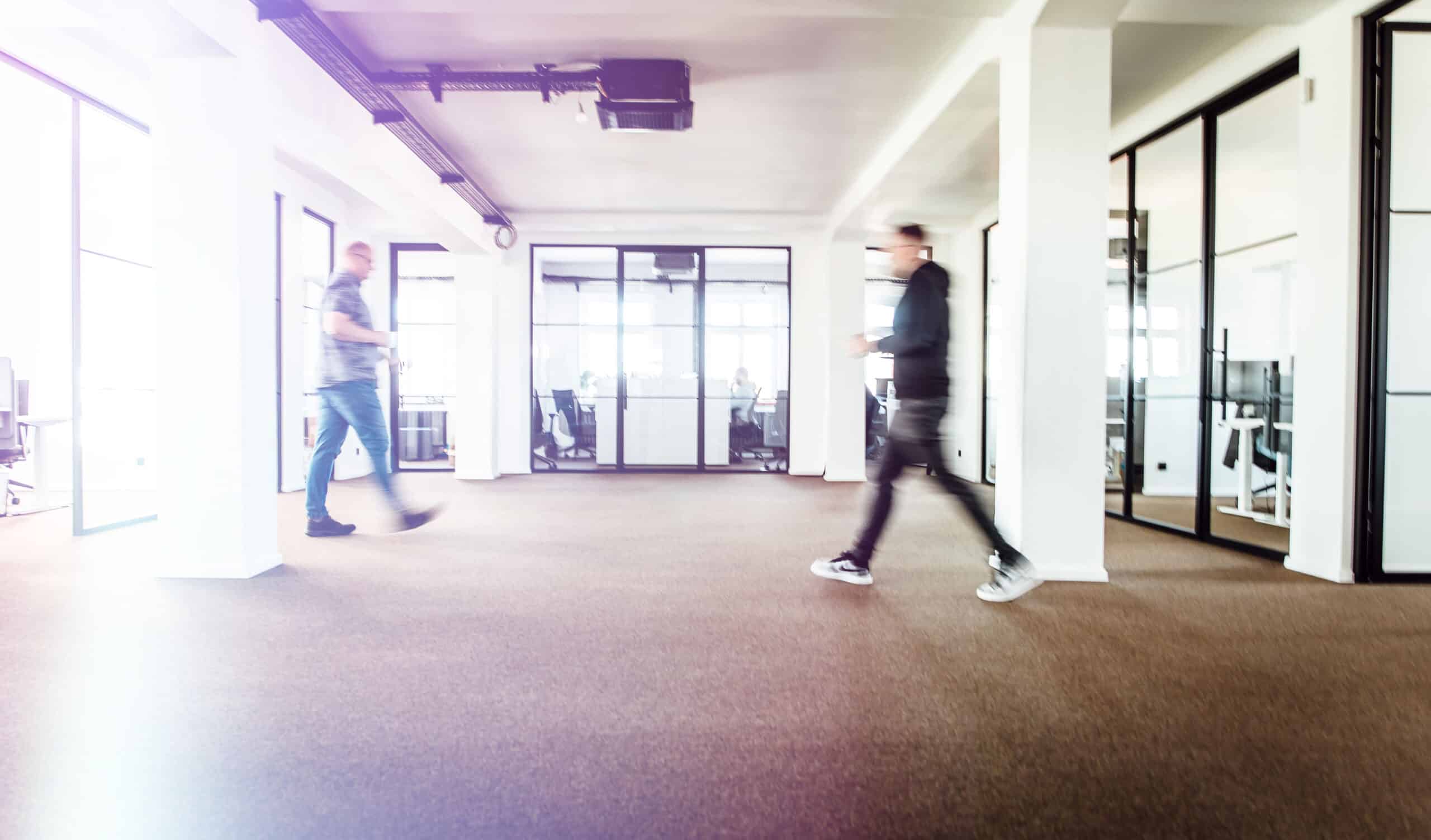 Zwei Personen gehen durch einen hellen, modernen Büroraum mit gläsernen Besprechungsräumen und Arbeitsplätzen im Hintergrund. Das Bild hat einen Bewegungsunschärfe-Effekt, der die Bewegung betont.