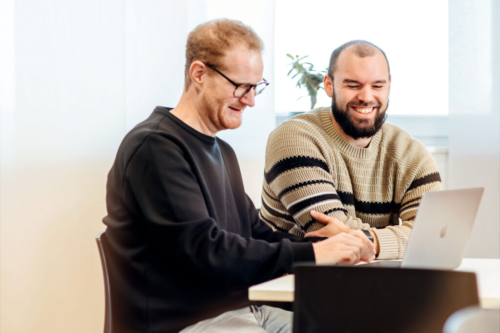 Zwei Männer sitzen lächelnd an einem Tisch und arbeiten gemeinsam an einem Laptop. Einer trägt eine Brille und ein dunkles Sweatshirt, der andere hat einen Bart und trägt einen gestreiften Pullover. Im Hintergrund ist eine Pflanze zu sehen.
