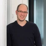 Ein Mann mit kurzen Haaren und Brille, der ein dunkles langärmeliges Hemd trägt, steht in einem Innenraum an einer weißen Wand und lächelt in die Kamera. Im Hintergrund sind Glasscheiben und ein schwach beleuchteter Flur zu sehen, an dem Menschen vorbeigehen.