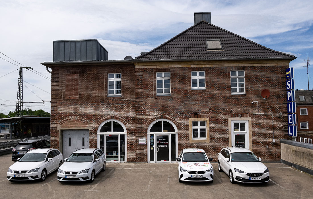 Ein Backsteingebäude mit drei davor geparkten weißen Autos und einem seitlich geparkten schwarzen Auto. Ein blaues vertikales SPIEL-Schild befindet sich auf der rechten Seite des Gebäudes. Auf der linken Seite sind ein Bahngleis und Stromleitungen zu sehen.