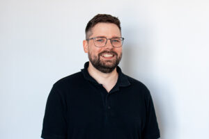 Ein Mann mit kurzen braunen Haaren, einem Bart und einer Brille steht lächelnd vor einem schlichten weißen Hintergrund. Er trägt ein schwarzes Polohemd, das den freundlichen und zugänglichen Geist einfängt, der oft in Menschen-Porträts zu sehen ist.