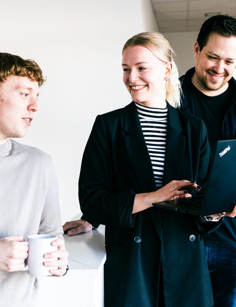 Drei Personen stehen in einem Innenraum zusammen. Eine Person hält einen Kaffeebecher in der Hand, eine andere hält einen Laptop und lächelt. Die dritte Person, ebenfalls lächelnd, schaut auf den Laptop, während sie ein freundliches Gespräch über ihre Ausbildung führen.