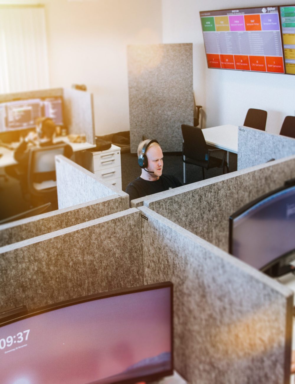 In einem modernen Büro eines Unternehmens arbeiten drei Personen an getrennten Arbeitsplätzen mit großen Monitoren. Zwei an der Wand befestigte Bildschirme zeigen Daten an, und der Arbeitsbereich ist durch ein Fenster mit natürlichem Licht erhellt.