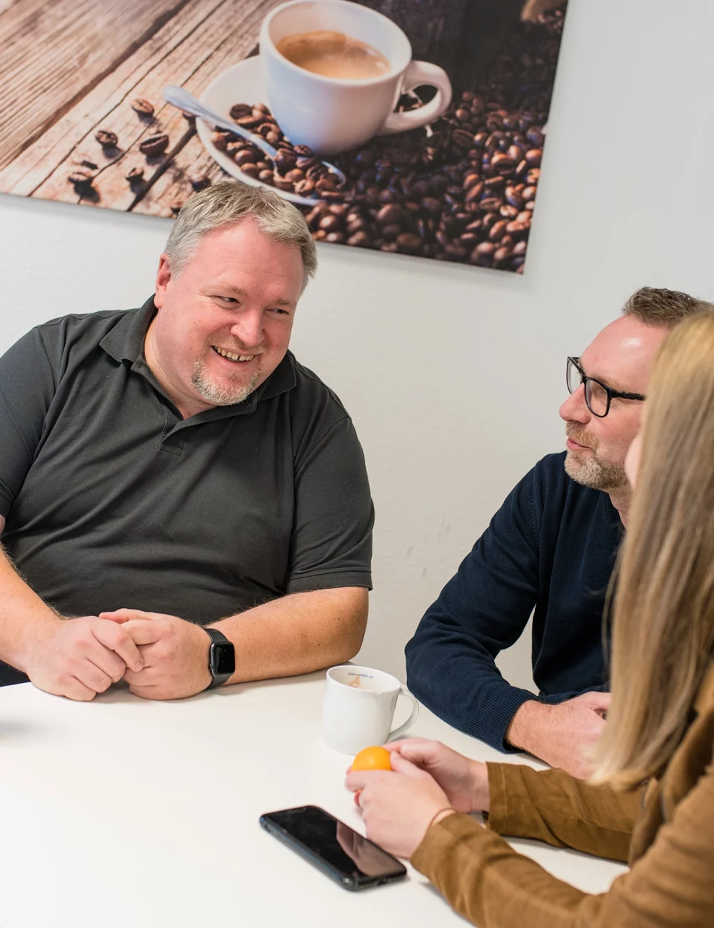 Drei Personen sitzen an einem kleinen Tisch und unterhalten sich angeregt über die Vorteile von Kaffee. Eine Kaffeetasse, eine Wasserflasche und ein Telefon stehen auf dem Tisch. Im Hintergrund hängt ein Wandbild mit einer Kaffeetasse und Bohnen.