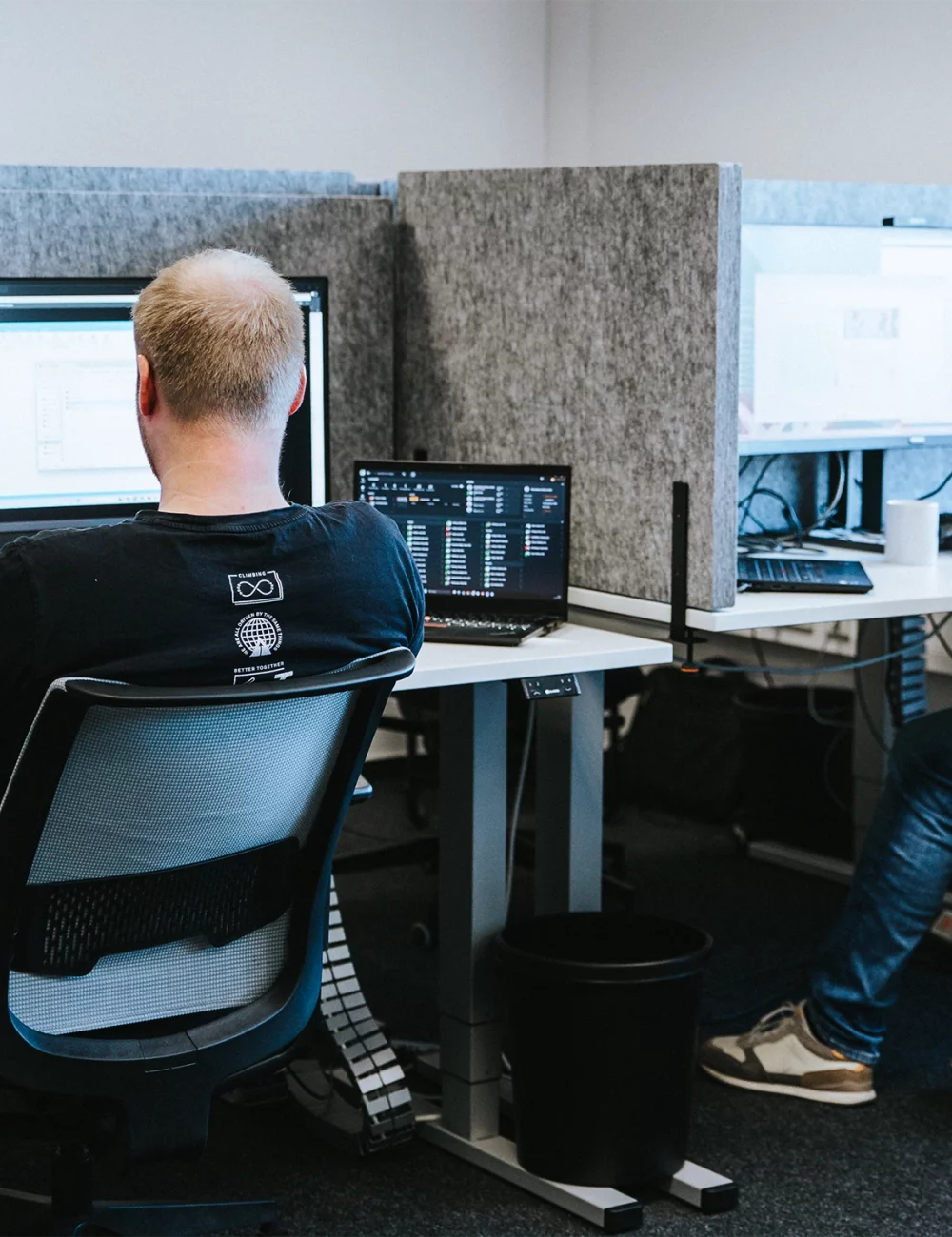 Zwei Männer arbeiten an Schreibtischen mit großen Monitoren in einem modernen Büro. Der eine blickt auf einen Bildschirm, auf dem Terra abgebildet ist und dessen Vorteile hervorgehoben werden, während der andere auf seiner Tastatur tippt. Beide sitzen auf Bürostühlen in einem Arbeitsbereich mit grauen Trennwänden.