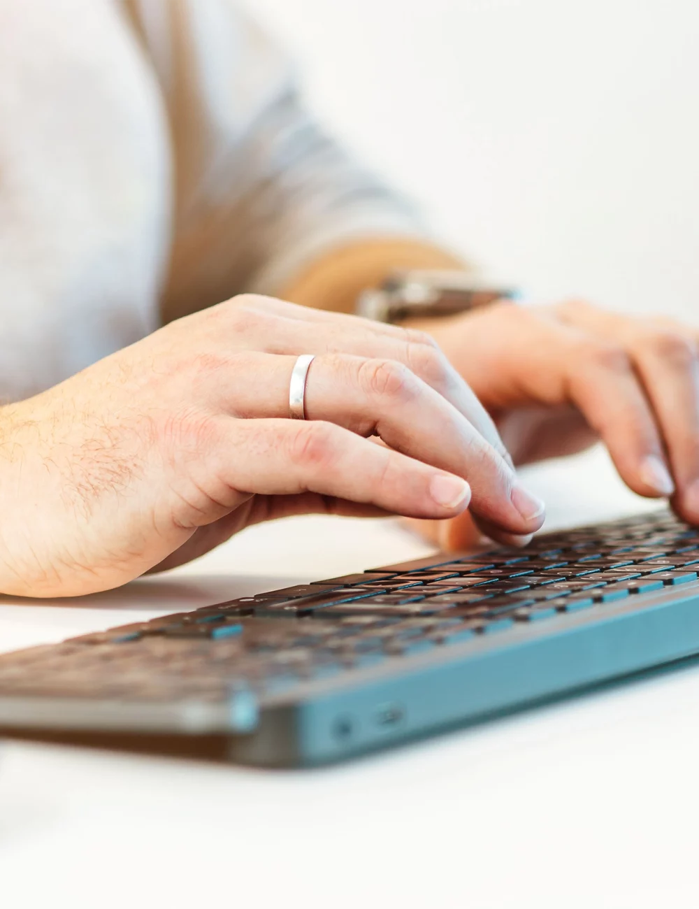Eine Nahaufnahme einer Person, die einen Ring an der linken Hand trägt und auf einer schwarzen Computertastatur tippt, während sie an einem weißen Schreibtisch sitzt, unterstreicht die Vorteile eines organisierten Arbeitsplatzes. Im Vordergrund ist eine Computermaus zu sehen.