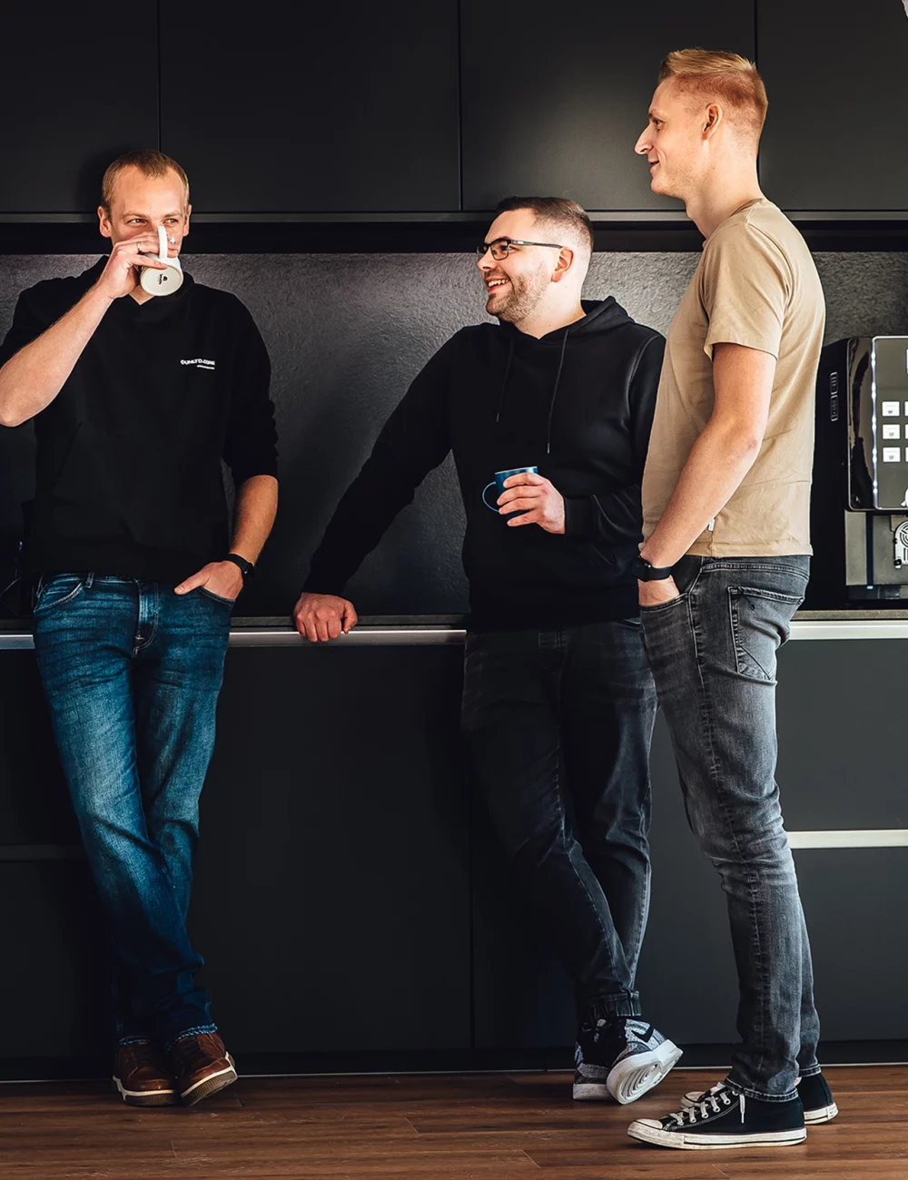 Vier Männer stehen und unterhalten sich lässig in einer modernen Emvion-Küche, halten Tassen und lächeln. Im Hintergrund sind dunkle Schränke, eine Kaffeemaschine und ein Wasserspender zu sehen.