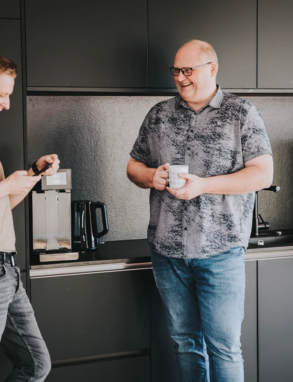Zwei Männer stehen in einer modernen Küche, der eine schaut auf sein Handy, der andere hält lächelnd eine Tasse in der Hand und genießt die Vorzüge eines gut ausgestatteten Raums mit einer Kaffeemaschine und einem Wasserkocher auf dem Tresen hinter ihnen.