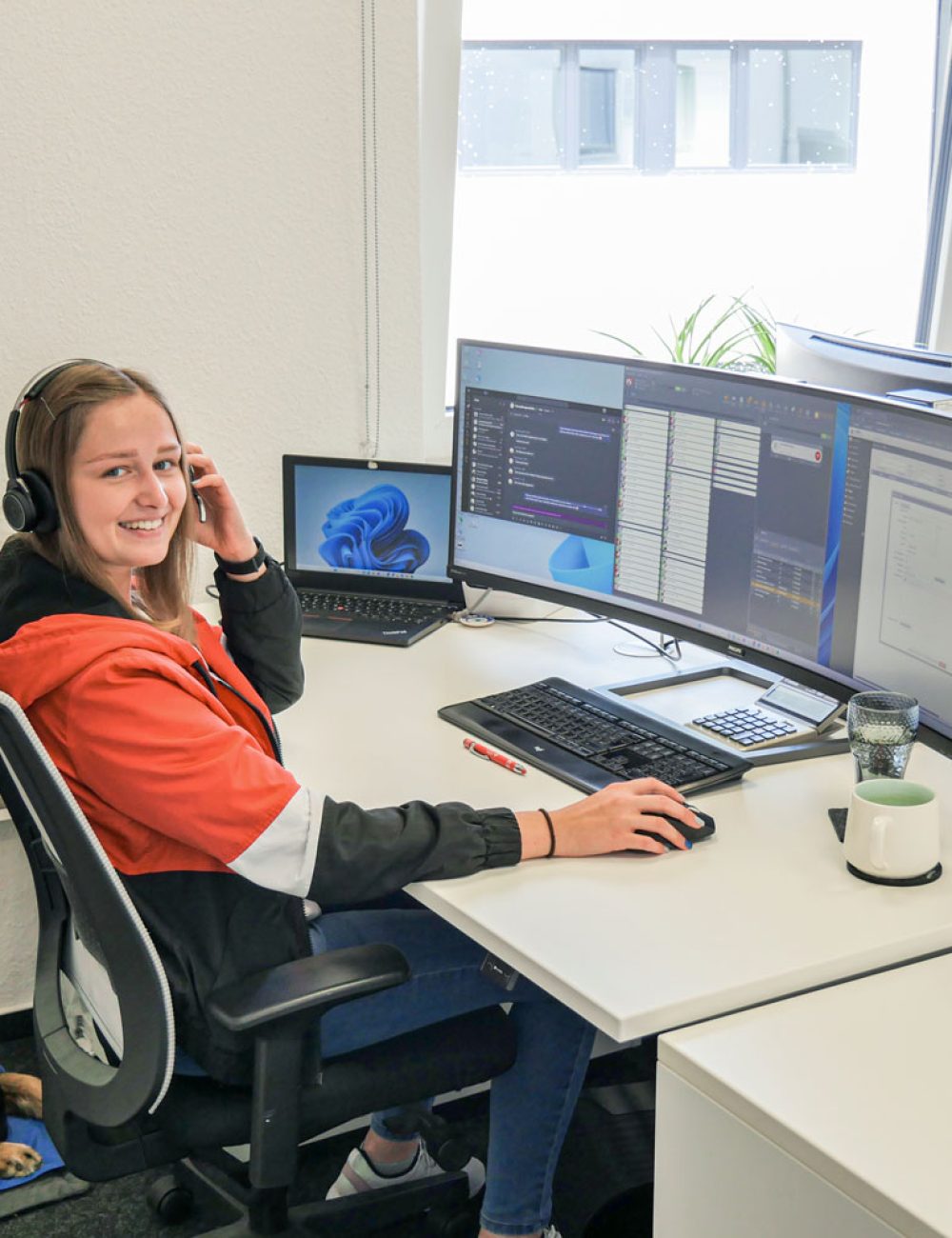 Eine Frau mit Kopfhörern und einer roten Jacke sitzt an einem von Bau & Handwerk inspirierten Schreibtisch mit zwei großen Monitoren und einem Laptop und lächelt in die Kamera. Ein Hund liegt auf einer Matte unter dem Schreibtisch. Auf dem Schreibtisch stehen ein Kaffeebecher und ein Glas.