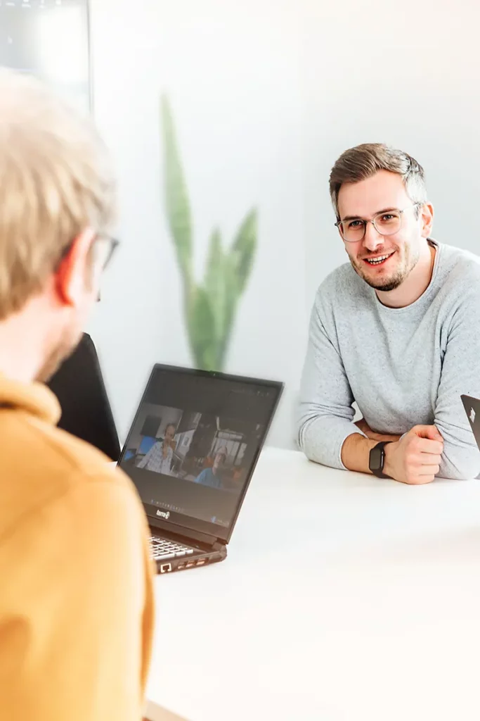 Zwei Männer unterhalten sich im Büro und vor ihnen sind ihre Laptops.