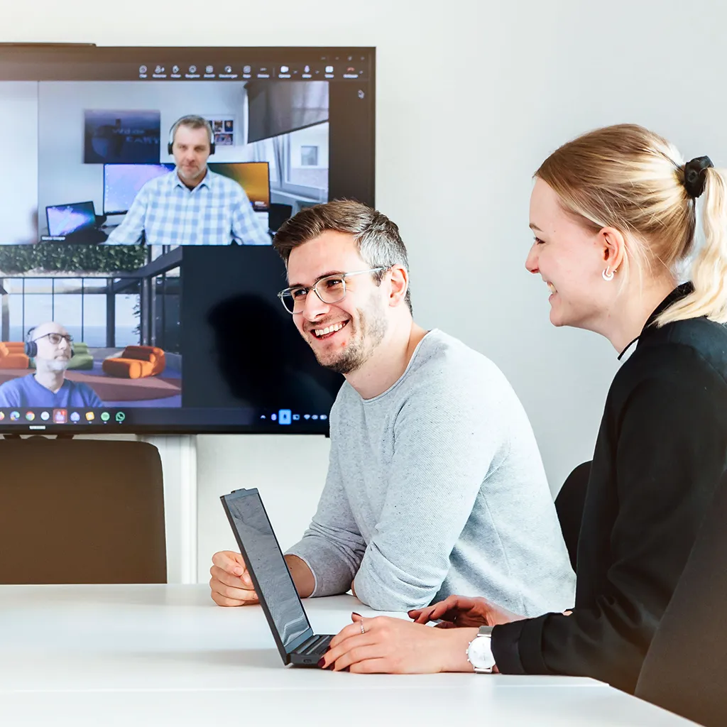 Zwei Personen sitzen während einer Videokonferenz im elektronischen Geschäftsverkehr lächelnd an einem Tisch, vor ihnen ein Laptop. Hinter ihnen zeigt ein Bildschirm drei Kollegen, die von verschiedenen Standorten aus an der Sitzung teilnehmen.