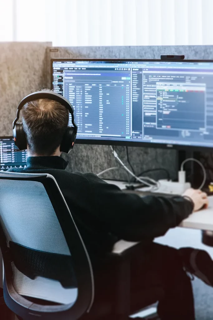 Eine Person mit Kopfhörern sitzt an einem Schreibtisch und blickt auf zwei große Computermonitore, auf denen Softwareentwicklungstools, Code und Reporting & BI-Dashboards angezeigt werden. Der moderne Arbeitsbereich ist gut beleuchtet und organisiert.
